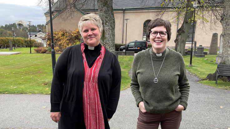 Två leende kvinnor står framför kyrka med kyrkogård.